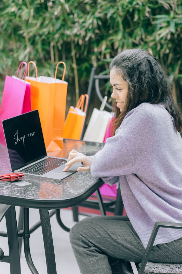 A woman shops online using a laptop outdoors, surrounded by colorful shopping bags, representing modern retail therapy.